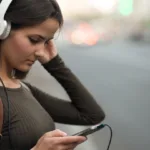 Woman with white headphones on looks down at her phone as she's about to cross a street.
