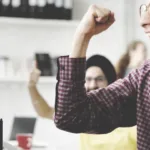 Man in glasses and checkered shirt raises his right fist in celebration while his mouth is open as if he's yelling something in excitement.