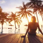 Silhouette of a person lounging on a pool deck with palm trees in front of them as the sun sets.