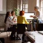 Three women sit in an open off around a table while a fourth sits on a window sill explaining something to the group.