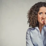 Woman in blue button-down top stands against a grey background chewing her thumbnail with a concerned look on her face. There are plenty of things new copywriters worry about that they don't need to worry about.
