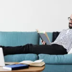 Man relaxing on blue couch holds a book with a laptop and books on table in front of him