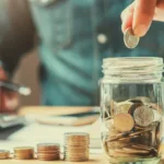 In blurred background is a torso in a denim shirt with a hand holding a pencil over a calculator, while in the focused foreground is a stack of quarters getting bigger as it goes toward a quarter-filled mason jar with a hand hovering over as it's about to drop a quarter.