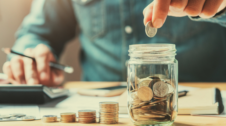 In blurred background is a torso in a denim shirt with a hand holding a pencil over a calculator, while in the focused foreground is a stack of quarters getting bigger as it goes toward a quarter-filled mason jar with a hand hovering over as it's about to drop a quarter.