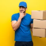 A man wearing a blue t-shirt and matching baseball cap with black shorts is standing against a yellow wall and holding three cardboard boxes. He also has a curious expression on his face.