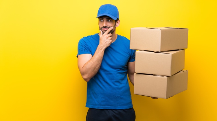 A man wearing a blue t-shirt and matching baseball cap with black shorts is standing against a yellow wall and holding three cardboard boxes. He also has a curious expression on his face.