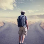 A man wearing a white fedora, blue shirt, and khaki shorts stands at a fork in the road in the middle of prairie land. He is carrying a navy backpack.