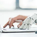 A robotic hand typing on a laptop next to a human hand also typing. The laptop is silver and the robotic hand is white.
