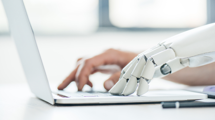 A robotic hand typing on a laptop next to a human hand also typing. The laptop is silver and the robotic hand is white.