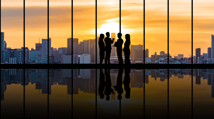 Four people are standing in an empty building in front of a large window. In the background there is a city skyline with the sun setting behind it.