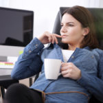 A woman is sitting in an office chair at a desk with a computer on it. She is wearing a jean jacket and holding a white coffee mug.