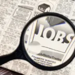 A magnifying glass is hovering over the classified section of a newspaper and through the lense there is a section of the paper that reads "jobs".