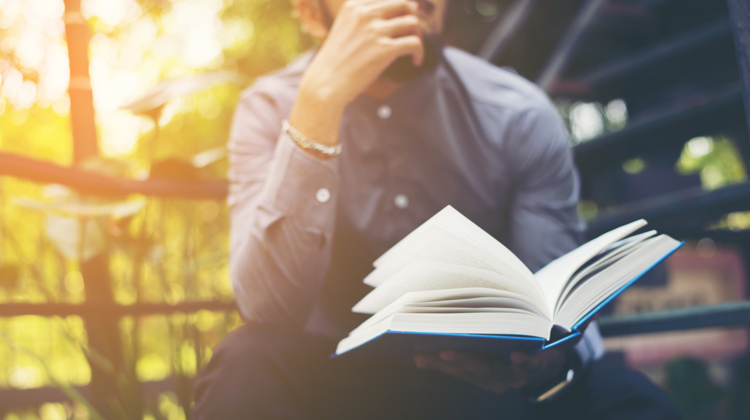 A man is sitting outdoors and the sun is beginning to set behind him. He is wearing a blue button up shirt and reading a book.