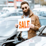 A man wearing sunglasses and a brown jacket is standing in the middle of a bunch of cars holding a sign that says "sale".