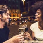 A man and a woman are touching their glasses of wine as to say "cheers". The man has white wine and the woman has red wine. They are sharing dinner outdoors in an urban setting.