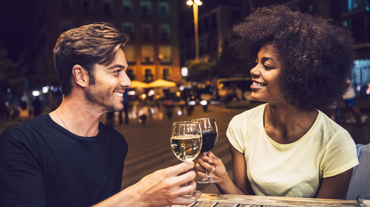 A man and a woman are touching their glasses of wine as to say "cheers". The man has white wine and the woman has red wine. They are sharing dinner outdoors in an urban setting.