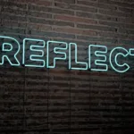 A neon sign that reads "reflect" that is mounted on a brick wall. The color of the sign is neon white.