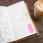A notebook sitting next to a cappuccino with a foam heart on top of it.