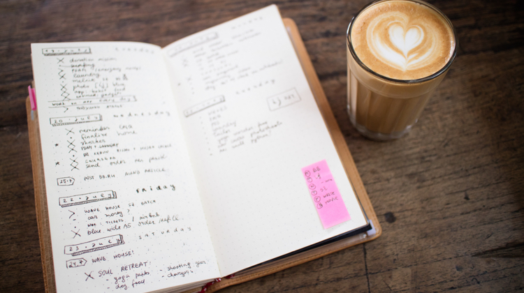 A notebook sitting next to a cappuccino with a foam heart on top of it.