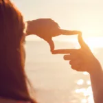 A woman looking at a sunset through an imaginary camera lense she is making with her fingers. The sun is reflecting off of the ocean water in front of her.