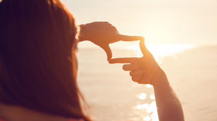 A woman looking at a sunset through an imaginary camera lense she is making with her fingers. The sun is reflecting off of the ocean water in front of her.
