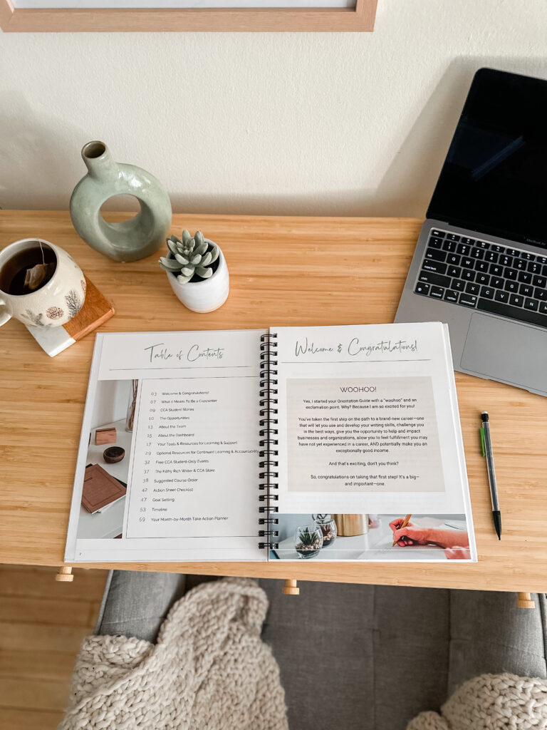 Image of the Comprehensive Copywriting Academy Orientation Guide on a desk next to a laptop, coffee mug, and succulent plant.