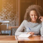 A woman wearing a striped long sleeve shirt taking notes while sitting at a table with a cup of coffee next to her. The room is lit softly.