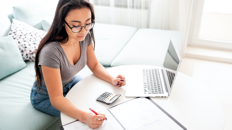 Woman with jeans and grey top with black rim glasses looks does at a calculator and papers with a red pen while her laptop is set to the side.