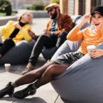 Woman in orange top and leather skirt and boots taps on her black newsboy cap while lounging in a beanbag chair with a coffee cup on a roof deck with two colleagues in the background.