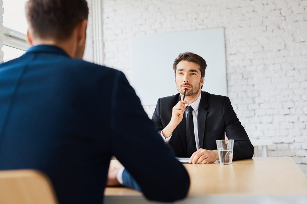 Man in black suit and black tie with white shirt sits across the negotiation table from a person in a blue suit jacket.