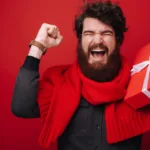 Bearded man holds up fist in excitement with mouth open in joy and red scarf while holding a red-wrapped gift in his other hand.
