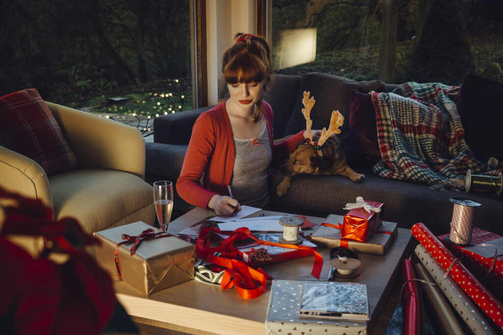Woman sits in front of a couch, where a dog sits with reindeer antlers, writing on a piece of paper while the coffee table in front of her is strewn with ribbon, gifts, and a glass of sparkling wine.