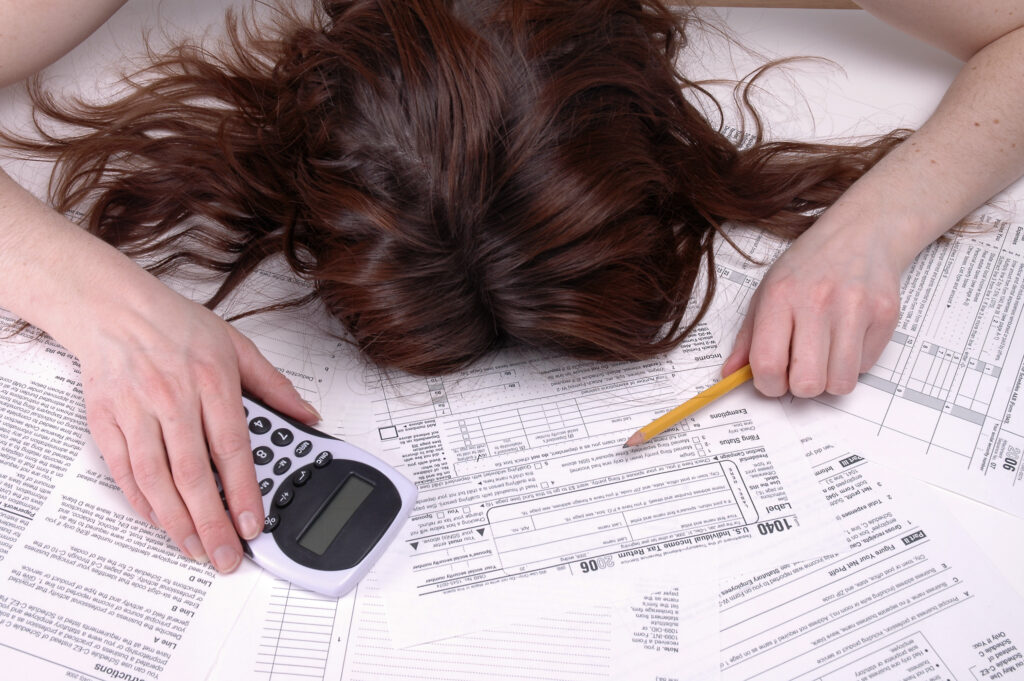 Woman is face down, head in a pile of tax documents with a calculator in one hand and a pencil in the other.