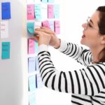 Woman in black-and-white striped shirt peels a teal sticky note from a sea of sticky notes in white, teal, and red, off a whiteboard.