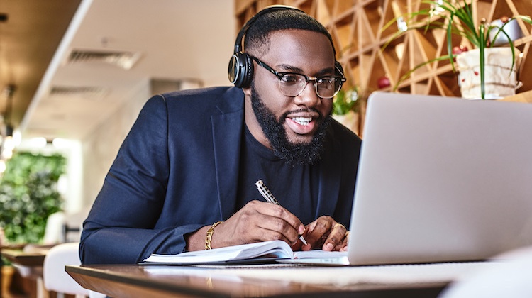 Black man in glasses, headphones, and blue blazer is in a cafe and eagerly writing down information while looking at his laptop.