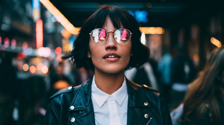 Woman with cropped haircut and leather jacket with white button-down top walks through a city, whose lights are reflected in her aviator glasses.