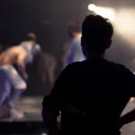 Silhouette of a man on stage with hands on his hips looking at performers in the spotlight.
