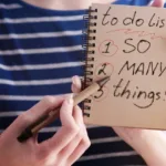 Person in blue shirt with white stripes points with a pen to a notebook that reads "to-do list: 1. so 2. many 3. things."