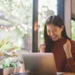 Young Asian woman sitting in front of laptop pumps both fists while smiling.