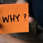 Hand holds out orange sticky note with the word "why" on it in marker.