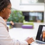 Woman in linen blazer smiles at her laptop screen where she is collaborating with another young woman via video conferencing.