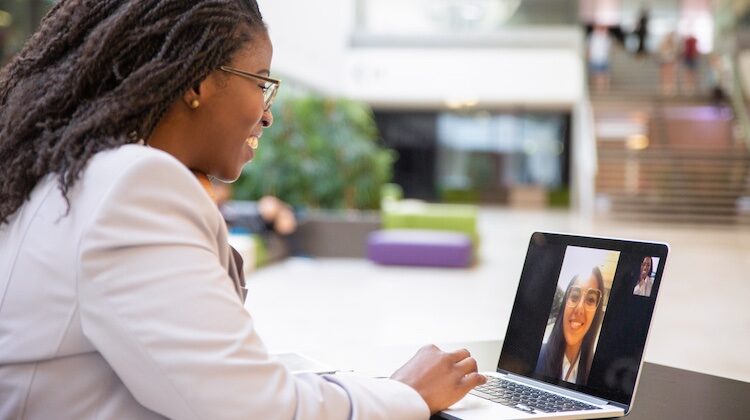 Woman in linen blazer smiles at her laptop screen where she is collaborating with another young woman via video conferencing.