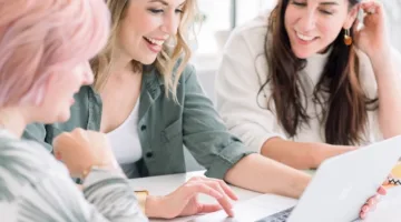 Learn about Filthy Rich Writer, including founder Nicki Krawczyk pictured in the middle here wearing a green shirt and scrolling on a laptop alongside two team members.