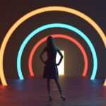 A woman wearing a dress stands in front of a door with neon rings of red, blue, and then yellow framing it.