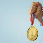 A forearm and fist against a light blue background tightly holds a gold medal with #1 on it with a red-white-and-blue-striped ribbon.