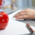 A Pomodoro timer sits on a notebook with a pen nearby and a person's hand in the background.