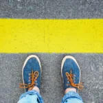 Two feet in blue shoes with leather laces stand on asphalt in front of a thick yellow line.
