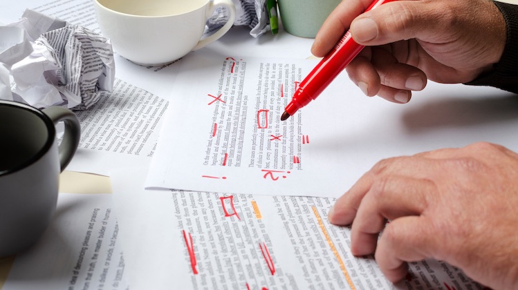 Two hands, the right one with a red marker, sit above two pieces of paper marking up the text.