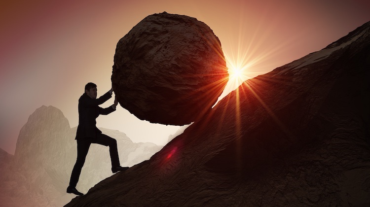 Man in suit is silhouetted against a setting sun as he scales a mountainside, pushing a large boulder up the mountain.