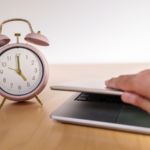 Hand lifts up a laptop that sits next to a pink old-fashioned alarm clock on a wooden desk.